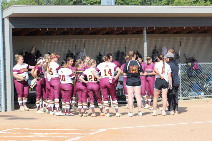A softball team in maroon uniforms gathers near the dugout for a meeting. Some players are in white jerseys, while others wear black team shirts. They stand in a circle, listening to a coach, with a fence and the dugout in the background.